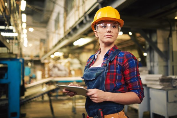 A female construction worker wearing a yellow hard hat, safety goggles, and holding a tablet, standing in a construction site, illustrating the use of Personal Protective Equipment (PPE).