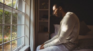 Side view of sad man sitting on bed by window