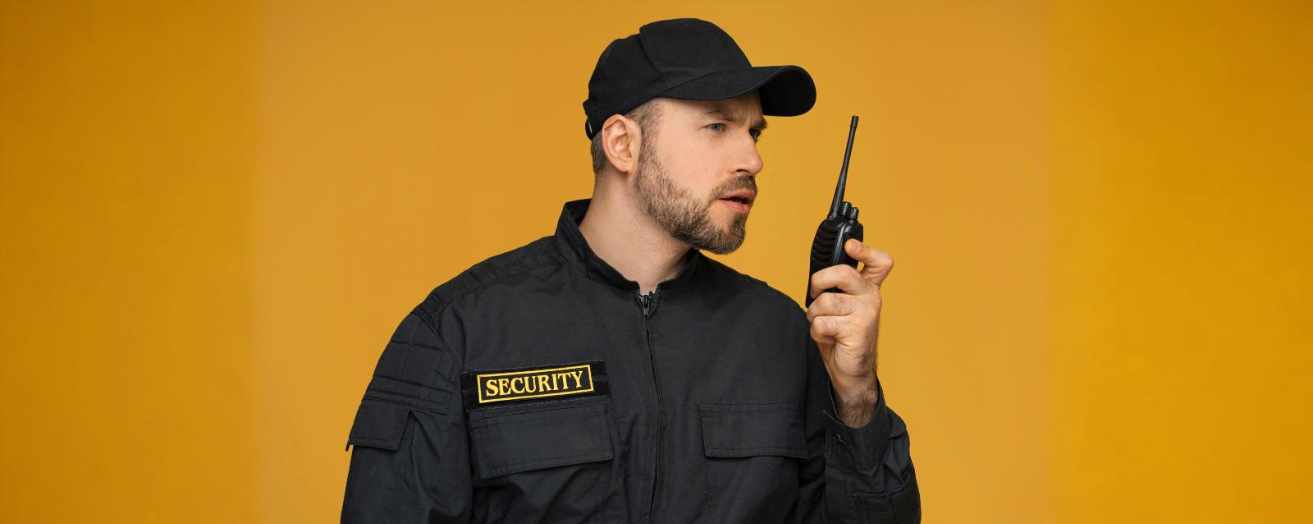 A professional security officer in uniform using a walkie-talkie, standing confidently against a bright yellow background, symbolising vigilance learned via door supervisor training.