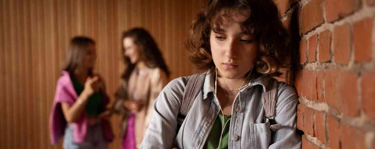 A teenage girl leans against a wall while two other girls whisper in the background, symbolising the isolation and vulnerability that can lead to grooming.